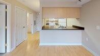 This single-floor unit offers a clean, functional layout designed for comfortable everyday living. The open-concept living and dining area is finished with light hardwood flooring that extends throughout the main living spaces, creating a warm and cohesive feel. Neutral beige walls and a textured ceiling complement the airy atmosphere, while a pendant light fixture adds a subtle decorative touch. A large window floods the living area with natural light and provides views of the surrounding environment.

The kitchen is positioned behind a half-wall peninsula that features a dark countertop, providing both additional prep space and a natural divider between the kitchen and living areas. Light wood cabinetry lines the upper portion of the kitchen, offering generous storage, while white appliances — including a range, over-the-range microwave, and refrigerator — complete the functional cooking space. Tile flooring defines the kitchen zone.

The bedroom is carpeted and generously proportioned, with a wide window overlooking a balcony railing and treetops beyond, bringing in natural light and a sense of connection to the outdoors. The full bathroom is finished with large-format beige tile flooring, a white vanity with ample cabinet storage, and a full soaking tub with shower — all presented in a clean, classic style.