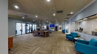 Located at 645 Ives Dairy Rd in Miami, FL, this single-story community facility serves as a well-designed clubhouse offering multiple functional areas under one roof. The entrance opens into a welcoming reception area finished with large-format tile flooring, soft gray walls, and decorative sunburst wall art above a rich wooden credenza, creating a polished first impression. A service window connects the lobby to a staffed back office, providing residents with convenient access to leasing staff through French-style interior doors.

The expansive main gathering area features patterned, multi-tone carpet tiles laid in a herringbone pattern, recessed lighting throughout a drop-panel ceiling, and soft blue-gray walls that lend the space a calm, cohesive aesthetic. This central room accommodates a large sectional sofa paired with a coffee table, a dining table with upholstered chairs, and vibrant teal accent seating arranged throughout. A wall-mounted flat-screen television serves as a media focal point.

A full glass wall with multi-panel black-framed doors provides direct visual and physical access to an on-site fitness center equipped with weight and strength training equipment. The open layout and abundant recessed lighting throughout create a bright, functional, and inviting environment suited for community gathering and daily activity.