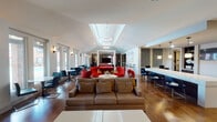 Located at 1700 Seaspray Ct in Houston, TX, this single-story community clubhouse is designed to offer a well-rounded social and recreational experience. The open-plan layout is anchored by a soaring vaulted ceiling with skylights that flood the central gathering area with natural light, complemented by striking circular pendant fixtures that add a modern touch.

The main lounge area features warm hardwood flooring and a mix of comfortable seating, including plush sofas and bold red accent chairs arranged around a central coffee table. A regulation-size billiards table with a vibrant red felt surface serves as a focal point for entertainment. Large windows and glass doors along the perimeter provide views of the surrounding outdoor areas, enhancing the airy ambiance throughout.

A long, sleek bar counter with blue upholstered bar stools is positioned adjacent to a kitchenette area, complete with pendant lighting, built-in shelving, a TV, and mosaic tile backsplash detailing. A separate lounge nook features contemporary black swivel chairs and a built-in desk, offering a quieter space for work or casual conversation.

The fitness center, visible through full glass walls, is well-equipped with cardio machines including ellipticals, treadmills, and stationary bikes, set beneath a coffered ceiling with recessed and ring lighting. Throughout the space, the design balances modern aesthetics with functional comfort.