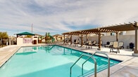 Located at 43808 Vintage St in Lancaster, CA, this single-level community amenity space is designed to serve as a central outdoor gathering hub for residents. At the heart of the area is a generously sized swimming pool with crystal-clear blue water, accessible via stainless steel entry rails and surrounded by a wide concrete pool deck. A substantial wood-beam pergola provides shaded relief from the Southern California sun, beneath which a row of cushioned lounge chairs and seating invites relaxation. Additional seating areas are distributed around the deck, offering ample space for sunbathing and socializing. Adjacent to the pool, a shallow wading area provides a gentler option for younger swimmers. A vibrant children's playground, equipped with a multi-level play structure and slide, sits just beyond the pool enclosure, making this space well-suited for families. The entire amenity area is enclosed by wrought iron security fencing with a gated entry, ensuring safety and controlled access. A posted rules board and waste receptacles reflect thoughtful facility management. Open skies and views of the surrounding single-story residential neighborhood complete the setting, giving the space a bright, community-oriented atmosphere.