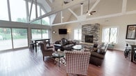 This single-story clubhouse is defined by its impressive architectural design, centered around a dramatic floor-to-ceiling stacked-stone fireplace that serves as the focal point of the main gathering area. The fireplace, finished with a clean stone hearth, is flanked by diamond-pane windows that frame outdoor views while flooding the space with natural light.

The vaulted ceiling rises dramatically overhead, accented by exposed white structural beams and ceiling fans, creating a sense of openness and grandeur. Rich hardwood flooring flows throughout the space, grounding the room with warmth and continuity.

The main lounge area features comfortable leather seating arranged around a glass-top coffee table, complemented by patterned accent chairs, creating an inviting and well-appointed gathering space. A wall-mounted television is positioned for easy viewing from the seating area.

A bar-height counter with a granite surface provides an additional gathering point, accompanied by sleek bar stools. Adjacent to this area, a dining space with high-top tables and seating offers views of the outdoors through large sliding glass doors. Decorative touches, including floral arrangements, mirrors, and wall art, contribute to the clubhouse's polished and welcoming ambiance.