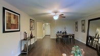 This single-floor residence offers a well-designed open-concept layout that seamlessly connects the living and dining areas, creating a spacious and inviting atmosphere. Rich, dark hardwood flooring runs throughout, providing a warm and elegant foundation that contrasts beautifully with the soft, neutral-toned walls.

The dining area serves as a focal point of the main living space, comfortably accommodating a full dining set with dark wood chairs and table, ideal for both everyday meals and entertaining. A ceiling fan with an integrated light fixture is centrally positioned overhead, providing both functional airflow and ambient lighting that enhances the room's warm, welcoming glow.

Decorative framed artwork adorns the walls, while a large statement mirror adds depth and visual interest to the space. A console table with tasteful décor accents one side of the room, contributing to the overall cohesive aesthetic.

A hallway extends from the main living area toward the entryway, where a solid front door provides a clear sense of entry and flow through the home. The layout is practical and open, allowing natural sightlines across the entire floor. The overall ambiance is warm, cozy, and well-appointed, making this space both functional and visually appealing.