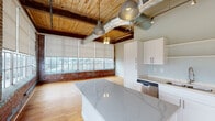 Located on E Main St in Haw River, NC, this single-floor industrial loft conversion masterfully balances historic character with modern updates. Original exposed brick walls, heavy timber beams, and tongue-and-groove wood-plank ceilings define the architectural identity of the space, while warm hardwood flooring runs throughout, tying each area together cohesively.

The open living area is generously proportioned, with banks of large, multi-pane industrial windows that flood the interior with natural light and frame views of the surrounding landscape. Exposed ductwork and ceiling fans add to the loft's industrial aesthetic.

The kitchen is a standout feature, equipped with white shaker-style cabinetry, a pale blue subway tile backsplash, and striking marble-veined quartz countertops — including a substantial central island. Stainless steel appliances, including a refrigerator, range, microwave, and dishwasher, complete the fully functional layout.

The bathrooms are finished with marble-style tile surrounds in the tub-shower combinations, quartz vanity tops, white cabinetry, and brushed nickel fixtures, delivering a clean and polished aesthetic. Additional rooms offer flexible space, each benefiting from the same hardwood flooring and abundant natural light. The overall ambiance is one of urban-industrial character thoughtfully refreshed with contemporary design sensibilities.