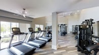 This fitness center offers a spacious, single-level layout designed to accommodate a full range of workout activities. The space is finished with light-toned, wood-style flooring that adds warmth while remaining practical and easy to maintain. Structural columns divide the floor plan into distinct zones without compromising the open feel of the room.

The cardio area is lined with a row of treadmills positioned along large windows, allowing natural light to fill the space and providing an airy, welcoming atmosphere. Ceiling fans are installed throughout, promoting air circulation and comfort during workouts. Recessed lighting supplements the natural light, ensuring the space remains well-illuminated at all hours.

Toward the rear of the fitness center, a dedicated strength training zone features a multi-station cable machine, free weights, and additional equipment stored on shelving units along the wall. Large mirrors are strategically placed to visually expand the space and allow users to monitor their form. The walls are finished in a neutral, warm gray tone that complements the overall clean and modern aesthetic, creating an environment that feels both functional and inviting.