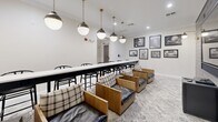 Located at 100 Harper's Crossing in Langhorne, Pennsylvania, this thoughtfully designed dining and lounge space blends modern aesthetics with comfortable, functional furnishings. The single-floor layout comprises a dining room and connecting hallway, offering a cohesive and inviting environment.

The dining room serves as the centerpiece, anchored by a long, sleek white communal table lined with classic black wishbone chairs. A row of statement globe pendant lights with brass fittings runs the length of the table, casting warm, focused light throughout. One wall features a dramatic dark shiplap panel, against which a flat-screen television is mounted, flanked by gold sconce lighting — creating a striking focal point. A curated gallery wall of black-and-white framed photographs adds a layer of character and visual interest.

Complementing the communal table, a cluster of solid wood lounge chairs with plaid and leather cushions provides a more relaxed seating option, set atop a soft patterned area rug. The hallway connects to a built-in wet bar niche, finished with the same dark shiplap backdrop and outfitted with a wine cooler and built-in microwave, ideal for entertaining.

Crisp white walls, crown molding, and light hardwood flooring in the hallway complete the refined, contemporary atmosphere of this well-appointed gathering space.