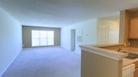 This single-floor unit presents an open-concept layout that seamlessly connects the kitchen and living areas. The living space is finished with wall-to-wall neutral-toned carpeting, creating a soft and cohesive feel throughout. A large window along the far wall allows natural light to fill the room, brightening the neutral beige and white color palette that defines the interior.

A kitchen peninsula extends into the space, providing a clear visual separation between the cooking and living zones while maintaining an open, airy flow. The peninsula features a light-toned countertop and offers practical utility for dining or food preparation.

The walls are painted in a warm neutral tone, complemented by crisp white trim and a textured ceiling. A wall-mounted HVAC vent is visible, indicating a built-in climate control system. Doorways lead to additional areas of the unit, suggesting access to further rooms beyond the main living space. The overall ambiance is clean, understated, and move-in ready, providing a blank canvas that accommodates a variety of interior design preferences.