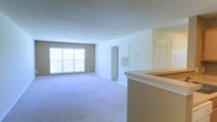 This single-floor unit presents an open-concept layout that seamlessly connects the kitchen and living areas. The living space is finished with wall-to-wall neutral-toned carpeting, creating a soft and cohesive feel throughout. A large window along the far wall allows natural light to fill the room, brightening the neutral beige and white color palette that defines the interior.

A kitchen peninsula extends into the space, providing a clear visual separation between the cooking and living zones while maintaining an open, airy flow. The peninsula features a light-toned countertop and offers practical utility for dining or food preparation.

The walls are painted in a warm neutral tone, complemented by crisp white trim and a textured ceiling. A wall-mounted HVAC vent is visible, indicating a built-in climate control system. Doorways lead to additional areas of the unit, suggesting access to further rooms beyond the main living space. The overall ambiance is clean, understated, and move-in ready, providing a blank canvas that accommodates a variety of interior design preferences.