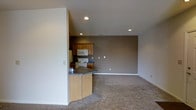 This single-floor residence at Glenmary Village offers an open-concept layout that combines the kitchen and living area into one cohesive space. The kitchen is outfitted with warm medium-toned wood cabinetry, a white electric range, over-the-range microwave, and a refrigerator. A peninsula with a dark countertop and light wood base provides a natural transition between the kitchen and the main living area, offering additional counter space and casual seating potential. The living area is generously sized, featuring wall-to-wall carpeting in a neutral beige tone that adds warmth underfoot. A taupe accent wall anchors the living space and adds depth to the otherwise light and airy color palette. Recessed lighting is positioned throughout the ceiling, providing even, consistent illumination across both the kitchen and living areas. The smooth white ceilings and clean white walls throughout contribute to a bright, open feel. The layout is practical and efficient, making the most of the available floor plan with a seamless flow between functional zones. Overall, the space presents a clean, move-in ready environment suited for comfortable everyday living.