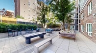Located at 4359 Roosevelt Way NE in Seattle's University District, this thoughtfully designed outdoor courtyard serves as a central amenity space for a multi-story residential community. The courtyard is finished with large-format light gray tile pavers throughout, creating a clean, modern aesthetic that complements the surrounding brick and painted-panel building facades.

The space is equipped with a slate-gray outdoor pool table positioned as a central gathering feature, alongside a life-sized chess set set into a dedicated tiled section of the courtyard. High-top bistro tables with blue chairs are scattered throughout, offering casual seating for residents. Concrete benches provide additional resting spots.

Lush greenery frames the perimeter, including mature trees that provide natural shade, ornamental shrubs, and planted stone-mulched beds that soften the hardscape. A raised landscaped terrace with a wooden privacy fence borders one side of the courtyard, adding depth and privacy. Wide concrete stairs with metal railings connect the courtyard to upper-level building access points and planted terraces.

The open-air design of the courtyard allows for abundant natural light throughout the day. The surrounding multi-story residential buildings with balconies overlook the space, creating an enclosed, community-oriented atmosphere ideal for social interaction and relaxation.