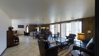 Located at 2311 West 16th Street in Greeley, Colorado, this single-floor space offers a welcoming and versatile environment designed for gathering and communal use. The open layout accommodates both casual lounging and dining, with clearly defined seating zones that flow seamlessly into one another.

The interior is anchored by a striking stone accent wall featuring tall, Gothic-style arched windows that lend the space a distinctive architectural character. These windows allow natural light to filter into the room, complementing the warm glow cast by pendant chandeliers suspended from the smooth, high ceiling above.

The dining area is furnished with multiple round and rectangular tables paired with dark wooden chairs, providing ample seating for groups. Adjacent to this, a comfortable lounge area features dark leather sofas and armchairs arranged around a wooden coffee table, accented by a richly patterned area rug that adds warmth to the light-toned tile flooring.

Along the exterior wall, a series of large sliding glass doors with vertical blinds flood the space with natural light and offer views to the outdoors. A wall-mounted television near the stone wall adds a modern, functional touch. Together, these elements create a multi-purpose space that is both functional and inviting.