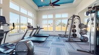 This fitness center is a thoughtfully designed, single-level workout facility that balances functionality with a bright, welcoming aesthetic. The space is defined by its elevated tray ceiling, finished in a bold teal accent that adds a vibrant pop of color against the crisp white walls, while a large ceiling fan ensures comfortable air circulation throughout. Multi-pane windows line the perimeter, flooding the interior with abundant natural light and offering open views of the surrounding outdoor environment, including greenery and adjacent buildings.

The flooring consists of modular carpet tiles in a mixed blue-grey palette, providing a clean and modern foundation underfoot. The fitness equipment is well-distributed across the space to allow for ease of movement. Cardio options include multiple treadmills, elliptical trainers, a stationary bike, and a rowing machine, all positioned to take advantage of the natural light from the large windows. A dedicated strength-training zone features a multi-use cable machine, a weight bench, a barbell rack with plates, and a full dumbbell rack. A large wall mirror expands the visual depth of the space and supports proper form during workouts, with a wall-mounted flat-screen television nearby. Access to an adjacent room is visible through a glass-paned sliding door.