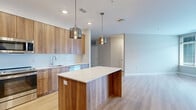 This well-appointed two-bedroom, two-bathroom unit offers a thoughtfully designed open-concept layout spread across a single primary floor. The kitchen serves as a central focal point, showcasing warm wood-tone cabinetry, white subway tile backsplash, white quartz countertops, and a full suite of stainless steel appliances including a refrigerator, range, and microwave. A large center island with matching wood-panel detailing provides additional workspace and seating, illuminated by two stylish pendant lights. Light wood-tone flooring flows throughout the main living areas, creating a clean, contemporary aesthetic.

The living area extends naturally from the kitchen, offering a versatile open space. Both bedrooms feature neutral tones and plush carpeting. The secondary bedroom benefits from a large corner window arrangement that invites ample natural light and urban courtyard views. Each bedroom is served by a dedicated closet with wire shelving systems for organized storage. The bathrooms are finished in neutral tile, with one featuring a glass-enclosed walk-in shower with white subway tile surround and chrome fixtures.

A private balcony accessible from the unit overlooks a well-maintained landscaped courtyard, offering pleasant outdoor views in a contemporary residential setting.