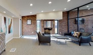 This single-floor community amenity space combines modern design with industrial-inspired architectural details to create a versatile and welcoming environment. Exposed brick accent walls serve as a defining feature throughout, adding warmth and texture that contrasts elegantly with the neutral carpeting, recessed lighting, and clean white walls.

The glass-enclosed fitness center is a standout element, enclosed by floor-to-ceiling black-framed glass walls that allow abundant natural light to flow inward while offering clear outdoor views of the surrounding landscape. The gym is equipped with a variety of cardio and strength-training machines, including treadmills, ellipticals, and cable equipment, with a mounted flat-screen TV and additional windows reinforcing the bright, airy atmosphere.

Adjacent to the fitness area, the main lounge space is generously sized and thoughtfully arranged. Leather seating, a round dining table with chairs, and a media console with a wall-mounted television create distinct zones for relaxation and socializing. A brick fireplace anchors one seating area, while colorful abstract artwork adds visual interest throughout.

A bar-height counter with stools connects to a kitchenette area, providing a convenient space for refreshments. Together, these areas form a cohesive, well-appointed amenity space designed for both activity and leisure.