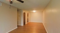 This single-story space presents a clean, open layout with warm hardwood-style flooring that extends throughout the main living area, contributing to a cohesive and inviting atmosphere. The neutral beige walls provide a versatile backdrop, complementing the natural tones of the flooring. A textured popcorn ceiling adds depth overhead, while a ceiling fan is positioned to support airflow and comfort throughout the space.

Flush-mount ceiling lighting provides consistent illumination across the living area. A floor vent along the baseboard indicates a central HVAC system for climate control. Clean white baseboards frame the perimeter of the room, adding a crisp finishing detail.

Toward the rear of the living area, a dark-stained interior door leads to an adjacent room, while a connected hallway or entryway reveals a glimpse of cabinetry, suggesting proximity to a kitchen or utility area. The overall layout appears straightforward and practical, with a clear sense of flow between spaces.

The space presents itself in a vacant state, making it easy to assess its proportions and imagine a variety of furniture arrangements within the open floor plan.