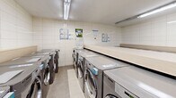 Located at 15 Eva Rd in Toronto's Etobicoke neighbourhood, this shared laundry facility offers a clean, well-organized space designed for practical, high-volume use. The room is finished with white ceramic wall tiles from floor to ceiling, lending a bright and hygienic atmosphere throughout. Fluorescent strip lighting runs along the ceiling, ensuring the entire space is well-illuminated.

Multiple front-loading LG washing machines and matching dryers are arranged in rows along the walls, providing ample capacity for building residents. Generous light wood-laminate folding tables are positioned centrally and along the perimeter, offering convenient surfaces for sorting and folding laundry. A large stainless steel utility sink with a wall-mounted faucet is available for hand-washing or pre-treating garments. A self-serve payment kiosk is conveniently located within the room, streamlining the payment process for machine use.

Informational notices and bulletin boards are mounted on the walls, keeping residents up to date on facility guidelines and building announcements. The neutral tile flooring is easy to maintain and complements the clean aesthetic of the space. A separate door provides access to an additional utility area. Overall, the facility is thoughtfully equipped to serve the everyday laundry needs of building residents efficiently and comfortably.