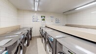 Located at 15 Eva Rd in Toronto's Etobicoke neighbourhood, this shared laundry facility offers a clean, well-organized space designed for practical, high-volume use. The room is finished with white ceramic wall tiles from floor to ceiling, lending a bright and hygienic atmosphere throughout. Fluorescent strip lighting runs along the ceiling, ensuring the entire space is well-illuminated.

Multiple front-loading LG washing machines and matching dryers are arranged in rows along the walls, providing ample capacity for building residents. Generous light wood-laminate folding tables are positioned centrally and along the perimeter, offering convenient surfaces for sorting and folding laundry. A large stainless steel utility sink with a wall-mounted faucet is available for hand-washing or pre-treating garments. A self-serve payment kiosk is conveniently located within the room, streamlining the payment process for machine use.

Informational notices and bulletin boards are mounted on the walls, keeping residents up to date on facility guidelines and building announcements. The neutral tile flooring is easy to maintain and complements the clean aesthetic of the space. A separate door provides access to an additional utility area. Overall, the facility is thoughtfully equipped to serve the everyday laundry needs of building residents efficiently and comfortably.