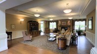 This generously proportioned single-floor common living space is thoughtfully designed to provide a comfortable and welcoming communal environment. The room features warm hardwood flooring throughout, anchored by a large floral area rug that defines the central seating arrangement. A standout stone fireplace with a wooden mantle serves as a natural focal point on one side of the room, flanked by decorative accents that enhance its inviting character. On the opposite wall, a substantial built-in wooden entertainment center houses a flat-screen television, providing a secondary gathering point for residents. The space is furnished with a mix of cream-toned sofas, upholstered armchairs, and a glass-topped coffee table, complemented by a distinctive cylindrical wooden side table. Pendant and dome-style ceiling light fixtures illuminate the room evenly, while large windows dressed with valances and drapes allow natural light to filter in, offering views of surrounding greenery. The drop ceiling with recessed panels adds a structured overhead element. Overall, the space conveys a warm, residential ambiance suited for relaxed socializing and leisure.
