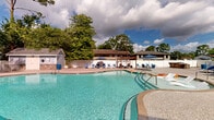 Located at 4955 Pepys Ln in Wilmington, NC, this two-floor property offers a well-appointed outdoor amenity area centered around a generously sized freeform swimming pool with crystal-clear turquoise water. The pool's curvilinear design creates an inviting resort-like atmosphere, complemented by a surrounding aggregate concrete deck that provides ample space for relaxation.

The outdoor area is thoughtfully furnished with numerous blue lounge chairs and patio umbrellas positioned throughout the sundeck, offering both sun and shade options for residents. A standout feature is a wooden pergola with a fabric canopy shade sail, beneath which comfortable seating with accent cushions and a round wicker coffee table create an intimate lounge setting.

The pool area is enclosed by a white metal fence and gate, providing security and privacy. A detached pool house structure with blue siding, stone accents, and a gabled roof serves as a utility and facilities building adjacent to the pool. Mature trees surround the perimeter, offering natural shade and a lush, green backdrop that enhances the outdoor ambiance.

The two-story residential building is visible in the background, featuring blue siding and multiple entry points with direct access to the pool deck, seamlessly connecting indoor and outdoor living spaces.