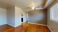 This single-floor space is characterized by its polished hardwood flooring, which flows consistently through the living areas and bedrooms, lending warmth and visual continuity throughout. The living room is a well-proportioned, open space anchored by a ceiling fan with a light fixture and neutral gray walls. A front entry door and window allow natural light to filter in, creating a welcoming atmosphere near the entrance.

The layout connects the living area to additional rooms through a central hallway. The kitchen features warm oak cabinetry, white laminate countertops, a stainless steel sink, and a black gas range with overhead ventilation—providing a functional and practical cooking space. Tile flooring in the kitchen contrasts with the hardwood found elsewhere.

The bedroom offers ample space, complete with a ceiling fan, a window overlooking greenery, and a built-in double closet with bifold doors, providing useful storage. The bathroom retains a retro character with sky-blue ceramic tile surrounding a full soaking tub, complemented by hexagonal floor tile and classic chrome fixtures.

Overall, the space presents a clean, straightforward layout with consistent finishes and a functional, comfortable ambiance throughout.