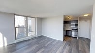 This single-floor urban residence offers a clean, contemporary layout designed to maximize both comfort and functionality. The open-concept living area is finished with wide-plank, hardwood-style flooring in a cool grey tone that lends a modern and cohesive aesthetic throughout the space. A large floor-to-ceiling window floods the living area with abundant natural light while framing elevated city views, creating a bright and airy atmosphere. A baseboard heating unit runs beneath the window, ensuring year-round comfort. The neutral white walls provide a versatile backdrop, complementing the understated yet polished design. Adjacent to the living area, the kitchen is efficiently appointed and features dark cabinetry paired with stainless steel appliances, including a refrigerator, gas range, built-in microwave, and dishwasher. A track lighting fixture overhead provides focused illumination across the workspace. The kitchen's warm wood-toned flooring creates a subtle distinction from the living area while maintaining visual harmony. The textured ceiling adds depth to the interior. Overall, the space presents a well-maintained, move-in ready environment with a practical urban design sensibility suited to modern living.