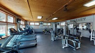 This fitness center offers a comprehensive and well-organized workout environment housed within a single-floor space characterized by a warm, wood-paneled ceiling and rubber flooring throughout. Multiple windows with wood-trimmed frames line the walls, allowing abundant natural light to fill the room while offering views of an adjacent outdoor pool area, creating an inviting and motivating atmosphere.

The cardio zone is clearly defined and features a row of treadmills, elliptical trainers, and stationary cycling bikes, providing a variety of options for cardiovascular exercise. The strength training area is equally well-appointed, with a multi-station cable and weight machine setup, a dedicated free weights section complete with a full dumbbell rack, barbells, and adjustable benches. A wall-mounted "WORKOUT" sign adds a motivational accent to the space.

Mirrors are strategically placed to enhance the sense of openness and assist with form during exercise. Ceiling fans ensure comfortable air circulation, supplemented by a wall-mounted HVAC unit. A mounted flat-screen television serves the cardio section. Additional functional training accessories, including resistance bands, a stability ball, and a floor exercise mat, round out the facility's offerings, making this a versatile and fully equipped space for a wide range of fitness activities.