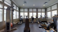 This fitness center is a thoughtfully designed single-floor facility built to support a comprehensive workout routine. Large windows line the walls, fitted with adjustable blinds that allow for controlled natural light, creating a bright and energizing atmosphere throughout the space. The dark rubber flooring provides a durable, slip-resistant surface well-suited for exercise and equipment use.

The cardio area is prominently featured and includes an impressive lineup of machines: elliptical trainers, upright and recumbent stationary bikes, and treadmills, all positioned to take advantage of the natural light and outdoor views. The equipment appears modern and well-maintained, with digital display consoles on select machines for tracking workout metrics.

Recessed ceiling lighting supplements the natural light, ensuring the space remains well-illuminated at all hours. The neutral, gray-toned wall color contributes to a clean, focused environment conducive to exercise. Additional fitness accessories, such as stability balls, are also present, offering options for supplemental training. The overall layout is open and organized, allowing for comfortable movement between equipment while maintaining an efficient use of the available floor space.