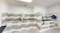 Located in Concord, NC, this purpose-built pet grooming room is a well-appointed, single-room facility designed with both functionality and aesthetics in mind. The space is immediately distinguished by its eye-catching tilework — a layered pattern of white subway tiles and sage green glass tiles that lines the walls, the built-in wash basin, and the grooming station surround, creating a clean and polished visual identity throughout.

The centerpiece of the room is a deep, tiled pet bathing tub equipped with a handheld showerhead and an accessible ramp, allowing pets of all sizes to enter comfortably. Adjacent to the tub is a raised grooming table with open shelving below and dual mounted pet dryers on either side, providing a complete wash-and-dry workflow in one compact area.

The flooring features large-format hexagonal tiles in a warm neutral tone, offering both durability and a refined finish. Bright, even lighting is provided by recessed ceiling fixtures and a skylight above the bathing area, ensuring excellent visibility throughout. A glass-paneled exterior door floods the room with natural light and provides convenient access to the outdoors. The bright white walls and clean lines of the space contribute to an overall atmosphere that is hygienic, modern, and welcoming for pet owners and their companions alike.