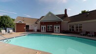 This clubhouse and pool area showcases a traditional architectural design, with a red brick exterior, neutral lap siding accents, a pitched shingle roof, and a prominent chimney. Multiple sets of white-framed French doors and large multi-pane windows line the building's facade, offering direct views of and access to the outdoor pool area. The generously sized outdoor pool features clear, blue-green water set within a wide concrete surround, ideal for relaxation and recreation.

Inside, the clubhouse interior is thoughtfully designed with an upscale, welcoming ambiance. The main lounge area is anchored by gleaming, dark polished tile floors and a coffered tray ceiling with recessed lighting and a ceiling fan, lending a sense of height and refinement. Elegant arched niches set into a stone-textured accent wall provide decorative display space, complementing the neutral color palette and rich wood crown molding throughout.

A seating area furnished with plush sofas and armchairs, as well as a separate dining and casual gathering space with glass-top tables, offer comfortable settings for socializing. Large windows frame direct views of the outdoor pool, filling the interior with natural light and creating a seamless connection between the indoor and outdoor spaces.