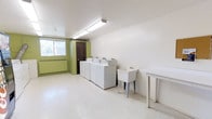This shared laundry facility is designed for practicality and ease of use. The room is well-lit by overhead fluorescent lighting, illuminating a clean, open layout with light-toned vinyl flooring that is easy to maintain. A row of white top-load washing machines lines one wall, providing ample capacity for residents or users. Adjacent to the machines sits a freestanding utility sink, ideal for pre-treating garments or handwashing delicate items. A sturdy white folding table is positioned nearby, offering a convenient surface for sorting and folding laundry. The walls are painted in a combination of white and a bold green accent, adding a touch of color to the functional space. A baseboard heater runs along the lower portion of the green-accented wall, ensuring comfort during colder months. Natural light filters in through a window on one side, complementing the overhead lighting. A dark wood door provides access to and from the room. The overall layout is straightforward and efficiently organized, prioritizing workflow and accessibility for laundry tasks.