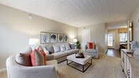 This two-story, three-bedroom home offers a thoughtfully designed layout spread across two well-proportioned floors. The main level features a welcoming living room adorned with a decorative textured ceiling, warm neutral tones, and plush carpeting. Natural light filters in through windows dressed with roman shades and blinds, creating a bright and comfortable gathering space. Adjacent to the living room, the kitchen is outfitted with dark cabinetry, black appliances including a range, microwave, and refrigerator, and durable hardwood-style flooring — providing a practical and efficient cooking environment. Upstairs, a hallway connects the three carpeted bedrooms, each finished in soft neutral tones. The generously sized primary bedroom includes a walk-in closet with built-in shelving, offering ample storage. The additional bedrooms each feature their own closets and are well-suited for a variety of uses. A staircase connects both floors seamlessly. Throughout the home, consistent use of warm beige and greige tones, textured ceilings, and plush carpeting creates a cohesive, comfortable ambiance, making this a practical and inviting multi-room residence.