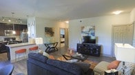 This single-floor residence features an open-concept living area designed for both comfort and functionality. The living room serves as the central hub of the space, anchored by a large, plush dark sectional sofa paired with a glass-top coffee table and coordinating accent furniture. Hardwood-style flooring runs throughout, adding warmth and visual continuity. A dark wood media console houses a flat-screen television and provides ample storage, while contemporary abstract artwork on the walls adds color and personality to the neutral-toned walls. Recessed lighting and a ceiling-mounted light fixture ensure the space is well-lit. A stylish console table with a sleek geometric base adds an additional decorative element. A partially visible corridor leads toward the bedroom, which can be glimpsed in the background and appears to feature similar finishes and natural light. The overall ambiance is modern and inviting, with a cohesive color palette of deep blues, warm oranges, and earthy tones lending the space a curated, contemporary aesthetic throughout.
