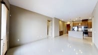 This single-floor space showcases a contemporary open-concept layout designed for both functionality and comfort. The living area is expansive, featuring large-format, high-gloss white tile flooring that reflects recessed ceiling lighting throughout, contributing to a bright and clean aesthetic. Neutral beige-toned walls provide a versatile backdrop, allowing the space to feel airy and welcoming.

The kitchen is integrated directly into the open floor plan and is distinguished by warm medium-toned wood cabinetry that contrasts attractively with the lighter surroundings. A white kitchen island anchors the space, fitted with a sink and served by pendant lighting overhead, making it a functional and visual centerpiece. Stainless steel appliances are visible along the kitchen wall, reinforcing the modern character of the space.

Recessed lighting is distributed throughout the ceiling, ensuring consistent illumination across all areas. A doorway visible from the main living area leads to additional rooms, suggesting a practical and well-organized floor plan. The overall ambiance is clean, modern, and understated, suited to a variety of interior design styles.