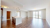Located at Harbor Point in Stamford, CT, this one-bedroom, one-bathroom residence offers a well-designed open-concept floor plan with generous living space. Rich hardwood flooring flows throughout the main living areas, anchoring the bright, airy aesthetic created by neutral white walls and abundant natural light.

The combined kitchen and living room serves as the heart of the home. The modern kitchen is outfitted with stainless steel appliances — including a gas range, built-in microwave, and full-size refrigerator — complemented by sleek, wood-grain cabinetry extending to the ceiling for ample storage. A large center island with a white countertop and undermount sink provides additional workspace and casual seating, with pendant lighting overhead.

The living area opens directly to a private balcony through sliding glass doors, offering views of the surrounding Stamford landscape. The spacious bedroom features plush carpeting, a large sliding glass door for natural light and outdoor access, and access to four closets, including a well-organized walk-in with built-in wire shelving.

The bathroom is finished with large-format tile flooring, a white vanity with modern cabinetry, and a full soaking tub with shower. The cohesive design throughout emphasizes clean lines, contemporary finishes, and functional living in a prime waterfront Stamford location.