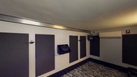 This single-floor studio space is purpose-built for professional audio recording or rehearsal, combining functional acoustic design with a polished, controlled environment. The room is lined with large, dark acoustic absorption panels strategically mounted across every wall, effectively minimizing unwanted sound reflections and ensuring optimal sonic conditions throughout the space. A patterned, textured carpet in deep blue and grey tones covers the floor, contributing to sound dampening while adding a refined aesthetic. Wall-mounted instrument hooks and dedicated music stand holders are positioned throughout the room, making it ready for immediate use by musicians or vocalists. A split-system air conditioning unit provides climate control without disrupting the acoustic integrity of the space. A large observation window connects the studio to an adjacent control room or monitoring area, allowing for clear sightlines between performers and engineers. Audio and signal routing equipment is mounted near the window for convenient connectivity. Accent lighting along the walls provides warm, focused illumination, creating a professional yet comfortable atmosphere for creative work. The space is accessed through a substantial door designed to maintain acoustic separation. Overall, this studio offers a well-equipped, thoughtfully designed environment for recording, rehearsal, or audio production.