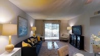 This single-story living space presents a thoughtfully designed and stylishly furnished interior. The living room serves as the focal point, featuring warm, neutral-toned walls that complement a carpeted floor and a comfortable, coordinated seating arrangement. A deep navy sofa adorned with yellow and patterned accent pillows anchors the space, paired with two geometric-print armchairs that add visual interest and texture.

A large sliding glass door dominates the far wall, allowing generous natural light to filter into the room while offering a direct view of a private outdoor patio area and surrounding greenery. Flanking the door, floor-length drapes in a warm tan tone frame the glass and contribute to the room's layered aesthetic.

A glass-topped coffee table with a metallic X-base sits centrally on the area rug, balancing elegance with practicality. Ambient lighting is provided by two table lamps positioned symmetrically on either side of the room, casting a warm glow. A black entertainment console with decorative lattice detailing houses a large flat-screen television. Wall art and carefully selected décor complete the polished, inviting atmosphere of this living space.
