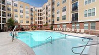 Located at 501 E Willard St in Durham, North Carolina, this single-level amenity space is part of a multi-story residential community defined by a classic combination of brick and neutral-toned siding. The centerpiece of the outdoor area is a generously sized resort-style swimming pool, enclosed within an open courtyard framed by the surrounding building. The pool area is lined with lounge chairs, providing ample space for relaxation under open skies. Adjacent to the pool, a wooden deck serves as an outdoor gathering area furnished with comfortable seating and accent chairs, complemented by landscaped planters, mature trees, and string lighting that lend a welcoming ambiance. A stone outdoor kitchen or grill station is also visible, enhancing the entertainment potential of the space. Entry into the interior is through glass-paneled doors that open directly to views of the pool and courtyard, maintaining a seamless indoor-outdoor connection. Inside, the lounge area features light hardwood-style flooring, tall ceilings, and large windows dressed with horizontal blinds that allow abundant natural light. A casual seating arrangement with a round table and modern chairs offers a relaxed setting, while a separate lounge nook adds additional comfort. The overall environment is bright, social, and thoughtfully designed for community living.