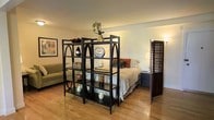 This thoughtfully designed single-floor space makes excellent use of an open-plan layout. The living and sleeping areas are elegantly separated by a striking dark-framed open shelving unit, featuring decorative arched details and multiple display tiers adorned with curated décor pieces. This functional divider provides a sense of definition between zones without sacrificing the open feel of the space. A wooden louvered room divider screen further enhances privacy near the sleeping area, adding a warm, layered aesthetic. The sleeping area is furnished with a neatly styled bed, complemented by a dark nightstand and accent lighting. On the living side, a plush olive-toned sofa sits against the wall alongside framed wall art, contributing to a cozy and inviting atmosphere. Throughout the space, warm-toned hardwood flooring adds richness and continuity, tying all elements together. The neutral, softly lit walls create a calm, welcoming ambiance. Overall, the space is characterized by its smart use of furnishings to organize an open floor plan, combining practicality with a tasteful, homey aesthetic.