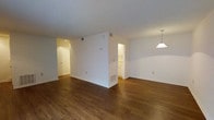 This 2-floor space features a clean, straightforward layout designed for comfortable everyday living. The main living area is open and expansive, finished with warm-toned hardwood-style flooring that extends throughout, creating a cohesive and inviting atmosphere. White walls and a textured ceiling keep the space bright and neutral, providing a versatile backdrop for a variety of interior styles.

A partial dividing wall separates the living area from the adjacent kitchen, which is visible through an open passageway. The kitchen features white cabinetry, offering ample storage, and benefits from the natural flow of light between rooms. A pendant light fixture hangs at the dining area edge, hinting at a dedicated space for meals.

A hallway on the opposite side of the living area leads to additional rooms, with a door visible at the far end suggesting private spaces such as bedrooms or utility areas. HVAC vents are positioned throughout, indicating a central climate control system. The overall ambiance is clean, airy, and move-in ready, with a neutral palette that allows residents to personalize the space with ease across both floors.
