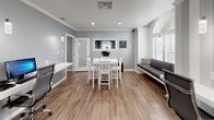This thoughtfully designed single-floor workspace blends modern aesthetics with functional layout, making it an ideal environment for focused work and collaboration. Rich wood-look flooring runs throughout, anchoring the space with warmth and continuity.

The main work area features a sleek, wall-mounted white desk with dual computer workstations, backed by a striking accent wall with bold vertical grey-and-white stripes. Pendant lighting drops elegantly above the workspace, providing focused illumination. Adjacent to the workstations, a contrasting dark wood-plank feature wall adds depth and texture, framing an archway that leads to the seating nook.

A built-in cushioned bench seat occupies a bright bay window area, fitted with plantation shutters that filter natural light beautifully. The arched transom window above adds architectural character and further enhances the brightness of the nook, creating an inviting spot to relax or collaborate informally.

The central area of the space accommodates a white high-top table with matching cross-back chairs, suitable for group meetings or collaborative tasks. A printer station and additional equipment are neatly integrated near the entrance. Crown molding trims the ceiling throughout, lending a polished, refined finish to the overall design. The soft grey walls tie all elements together into a cohesive, professional atmosphere.