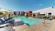 Located in Goodyear, Arizona, this outdoor amenity space showcases a resort-style pool area designed for relaxation and recreation under the abundant Arizona sunshine. The centerpiece is a large, sparkling swimming pool with crystal-clear turquoise water, complemented by a decorative water fountain feature that adds a dynamic visual element to the space.

Surrounding the pool is a generously sized concrete sun deck furnished with multiple blue lounge chairs and patio seating, providing ample space for sunbathing and outdoor leisure. Several large white patio umbrellas are strategically positioned throughout the deck, offering shaded retreats from the desert sun. Lush desert-friendly landscaping, including agave plants, palm trees, and ornamental shrubs, frames the area and adds greenery to the hardscape.

The pool area is enclosed by a safety fence and is flanked by the multi-story residential buildings, which feature a modern Southwestern architectural style with stucco exteriors in warm earth tones, accented by bold rust and deep red façade panels. A covered pergola structure adjacent to the pool provides an additional shaded gathering space. The overall ambiance is bright, open, and inviting, capturing the essence of an upscale outdoor lifestyle tailored to the warm Goodyear climate.
