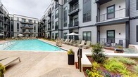 Located at 327–349 W Tremont Avenue in Charlotte's vibrant South End neighborhood, this property showcases a thoughtfully designed outdoor amenity level that serves as the centerpiece of the community. The courtyard is anchored by a generously sized resort-style swimming pool with clear blue water, surrounded by a spacious concrete pool deck furnished with rows of chaise lounges, shaded umbrella seating, and a covered grill station — ideal for both relaxation and social gatherings. Lush landscaping with seasonal flowers, ornamental grasses, and manicured garden beds lines the walkways, adding color and warmth to the hardscaped surroundings. A dedicated outdoor patio area features bistro-style tables and chairs nestled alongside the building's facade, offering a quieter setting for dining or casual conversation. The multi-story building exterior is clad in gray horizontal siding accented with dark trim, large windows, and private balconies that overlook the courtyard below. Glass entry doors branded with the Penrose name provide a polished arrival experience. The overall ambiance strikes a balance between contemporary urban design and inviting outdoor living, creating a cohesive and well-appointed community amenity space.