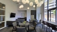 This stylish single-floor communal space is designed with a contemporary aesthetic that seamlessly blends functionality and modern elegance. The room immediately draws the eye upward to its soaring ceilings, from which a striking cluster of spherical white globe pendant lights descend at varying heights, creating a dramatic and visually captivating focal point.

The seating area is thoughtfully arranged with a mix of plush gray sofas and sleek dark armchairs grouped around round tables, offering flexible arrangements suited for both casual conversation and small gatherings. Fresh floral accents on the tables add a welcoming touch of color against the neutral palette of grays and blacks throughout.

A wall-mounted flat-screen television anchors one side of the room, adding to the lounge's entertainment appeal. Adjacent to the seating area, a compact kitchenette provides practical amenities including a full-size stainless steel refrigerator and microwave, supported by dark cabinetry that reinforces the space's modern tone.

Large windows and a glass-paneled exterior door allow abundant natural light to flood the interior, connecting the space to the outdoors and enhancing the open, airy atmosphere. The combination of high ceilings, curated lighting, and refined furnishings makes this lounge an inviting and well-appointed communal environment.