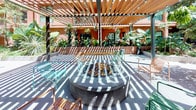 This thoughtfully designed outdoor courtyard serves as a central communal amenity space set within a multi-story residential building. A striking wood-slat pergola with suspended string lights spans the gathering area, casting dynamic patterns of light and shadow across the composite deck below. The courtyard is organized into distinct functional zones, including a central circular fire pit surrounded by a glass windscreen and colorful modern seating, ideal for relaxed social gatherings. A dedicated outdoor kitchen area features stainless steel BBQ grills and prep surfaces, accommodating al fresco dining and entertaining. Lush, densely planted raised garden beds line the walkways and perimeter, filled with a rich variety of tropical and desert plants including monstera, bromeliads, succulents, cacti, and palms, creating a verdant, resort-like atmosphere. Wide, smooth pathways connect the various zones and encourage easy movement throughout the space. The warm terracotta tones of the surrounding building facades complement the natural wood and greenery, giving the courtyard a cohesive, inviting aesthetic. Abundant natural light filters through the pergola, enhancing the open-air ambiance of this well-appointed outdoor living space.