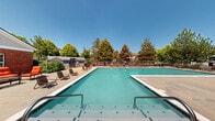 Located at 630 S Peck Dr in Longmont, Colorado, this outdoor pool facility is a well-appointed communal amenity space designed for relaxation and recreation. The centerpiece is a generously sized swimming pool with clear turquoise water, accessible via wide entry steps with stainless steel handrails and marked with decorative tile accents along the pool floor. The expansive surrounding deck provides ample room for lounging, furnished with a variety of chaise lounges, chairs, and seating groupings arranged throughout the patio area. Shaded umbrella tables and vibrant outdoor sofas offer comfortable spots for gathering and socializing. A covered outdoor kitchen station is prominently featured, equipped with a large stainless steel grill and additional cooking appliances, making it ideal for outdoor dining and entertaining. The facility is set against a backdrop of mature trees that provide natural shade and privacy, while the two-story brick and siding clubhouse building serves as an attractive architectural anchor to the space. The entire area is enclosed by a secure wrought iron fence, and the open Colorado sky above ensures the space is bathed in abundant natural light throughout the day.