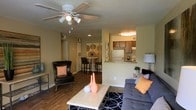 This single-floor space showcases an open-concept design that seamlessly connects the living room, dining area, and kitchen. The living room is anchored by a plush gray sofa accented with orange throw pillows, complemented by a cushioned armchair and a light-toned coffee table resting atop a bold patterned area rug. Warm wood-style flooring runs throughout, adding continuity and a welcoming feel to the space. A ceiling fan with integrated lighting fixture provides both airflow and ambient illumination overhead.

Adjacent to the living area, a cozy dining nook features a bistro-style high table paired with bar stools, ideal for casual meals. The kitchen is positioned at the rear and is accessible via a pass-through counter that also functions as a breakfast bar. It features dark wood cabinetry, a white refrigerator, and a sink positioned beneath overhead cabinet storage. Warm overhead lighting in the kitchen creates an inviting ambiance. Neutral beige walls with subtle decorative accents tie the overall aesthetic together, giving the space a cohesive and comfortable character throughout.