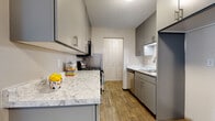 This single-floor residence offers a thoughtfully laid-out interior that balances functionality with modern updates. The galley-style kitchen is a standout feature, showcasing sleek gray cabinetry, marble-look countertops, wood-look luxury vinyl flooring, a gas range, a dishwasher, and a stainless sink — all beneath a textured ceiling. A set of louvered bi-fold closet doors provides convenient pantry or utility storage just off the kitchen. The open living and dining area flows seamlessly, with plush carpeting in the living space transitioning to wood-look flooring in the dining nook. The dining area comfortably accommodates a round table and chairs, with textured walls adding subtle character. The living room benefits from a large sliding glass window that invites natural light and offers views of mature greenery outside. The bedroom is generously sized, finished with soft carpet, a ceiling fan with light fixture, a baseboard heater, and a wide window framing outdoor tree views. The full bathroom features classic white subway tile surrounding a soaking tub and shower combination, paired with brushed nickel fixtures and a clean, bright finish throughout.
