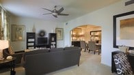 This single-floor residence offers a thoughtfully designed layout that balances comfort and style. The living area serves as the heart of the home, featuring light-toned tile flooring, crisp white walls, and a modern ceiling fan that adds both function and a contemporary touch. A sleek dark entertainment unit anchors the room, complemented by coordinating dark gray sofas and curated decorative accents that lend the space a refined, cohesive aesthetic. A striking arched passageway draws the eye from the living area into the dining room beyond, creating a seamless visual and physical connection between the two spaces. The dining room is warmly illuminated by a pair of elegant table lamps, casting a rich, inviting glow over the dark wood dining table and patterned upholstered chairs. A dark sideboard and decorative mirror complete the room's sophisticated look. Throughout the space, the interplay of warm lighting, neutral tones, and dark furnishings creates a balanced ambiance that feels both stylish and welcoming. The open-concept flow between the living and dining areas enhances the sense of spaciousness, making this residence well-suited for both everyday living and entertaining.