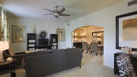 This single-floor residence offers a thoughtfully designed layout that balances comfort and style. The living area serves as the heart of the home, featuring light-toned tile flooring, crisp white walls, and a modern ceiling fan that adds both function and a contemporary touch. A sleek dark entertainment unit anchors the room, complemented by coordinating dark gray sofas and curated decorative accents that lend the space a refined, cohesive aesthetic. A striking arched passageway draws the eye from the living area into the dining room beyond, creating a seamless visual and physical connection between the two spaces. The dining room is warmly illuminated by a pair of elegant table lamps, casting a rich, inviting glow over the dark wood dining table and patterned upholstered chairs. A dark sideboard and decorative mirror complete the room's sophisticated look. Throughout the space, the interplay of warm lighting, neutral tones, and dark furnishings creates a balanced ambiance that feels both stylish and welcoming. The open-concept flow between the living and dining areas enhances the sense of spaciousness, making this residence well-suited for both everyday living and entertaining.