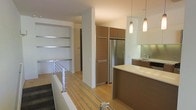 This contemporary two-floor space showcases a thoughtful blend of modern design and functional living. The open-plan kitchen is a standout feature, boasting warm wood-toned cabinetry that extends to the ceiling, stainless steel appliances including a full-size refrigerator, and a generous kitchen island with a clean white countertop. Pendant lighting hangs above the island, casting a warm, inviting glow over the space. The kitchen also features a dedicated sink area with built-in cabinetry and under-counter appliances, providing ample storage and workspace.

The interior is finished with light hardwood flooring throughout, contributing to a bright and airy atmosphere. A modern staircase with sleek metal horizontal railings connects the two floors, adding an architectural element that complements the overall contemporary aesthetic. At the upper level, built-in shelving units are integrated into the wall, offering both practical storage and display space.

The layout is open and spacious, with clean lines and a neutral color palette of whites and warm wood tones creating a cohesive and refined ambiance throughout. The design prioritizes both style and practicality, making this a well-appointed multi-level space.