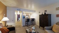 This single-story residence is designed with an open-concept layout that integrates the living area and kitchen into one cohesive space. The living area features wall-to-wall carpeting in a neutral tone, creating a warm and comfortable atmosphere. A coffee table serves as a central focal point, complemented by surrounding seating and a dark wood armoire that adds a traditional accent to the room.

The kitchen is positioned at the rear of the open space, elevated slightly behind a raised breakfast bar with dark bar stools providing casual seating. Pendant lighting hangs above the bar, illuminating the counter while contributing a modern touch to the overall design. The kitchen is equipped with appliances and cabinetry in light tones, providing a clean, functional workspace.

A hallway extending from the living area leads to the bedroom, which is visible in the background and features a distinctive accent wall in a deep plum tone, adding a bold design element. The textured ceiling throughout the space adds subtle visual depth, while ceiling vents indicate a central HVAC system. The overall ambiance is cozy and practical, with a cohesive neutral color palette punctuated by intentional design details.